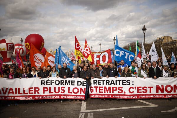 A crowd marches in Paris behind a long horizontal banner with black and red lettering on a white background protesting the pension overhaul. The demonstrators are also carrying other banners and flags with trade union logos in multiple colors.